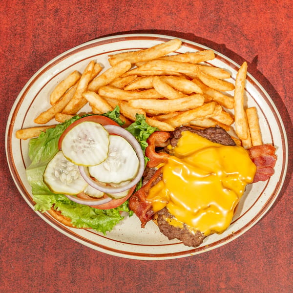 Overhead view of an open-faced cheeseburger with bacon, lettuce, tomato, pickles, and onions, served with fries.
